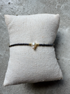 Armbånd med sorte perler og guld detaljer, centreret af en unik perle, på en lys pude.