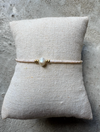 Beige perlearmbånd med central perle og gulddetaljer på tekstureret pude.